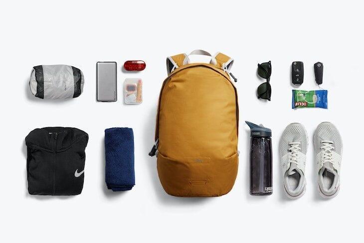 Gelber Bellroy Lite Daypack Rucksack mit Alltagsgegenständen wie Wasserflasche, Schuhe, Sonnenbrille, Handy, Handtuch und Jacke, flach angeordnet auf weißem Hintergrund.