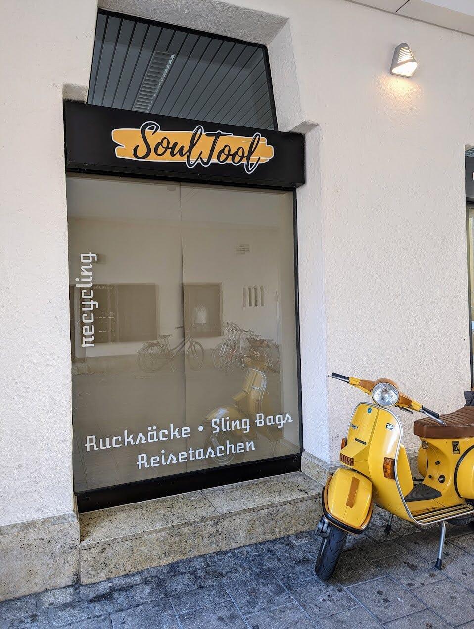 SoulTool Ladenfront mit Schaufenster, gelber Roller davor, Text: Rucksäcke, Sling Bags, Reisetaschen